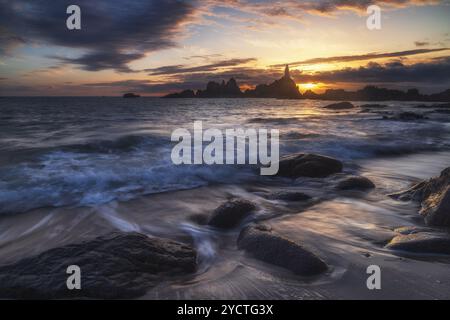 Leuchtturm Le Corbiere bei Sonnenuntergang bei Ebbe. Wellen und Steine am Strand. Stockfoto