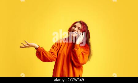 Lächelnde junge rothaarige Frau, die fröhlich auf dem Handy spricht und Gesten macht, gegen hellgelbe Rückwunde. Kommunikation. Stockfoto