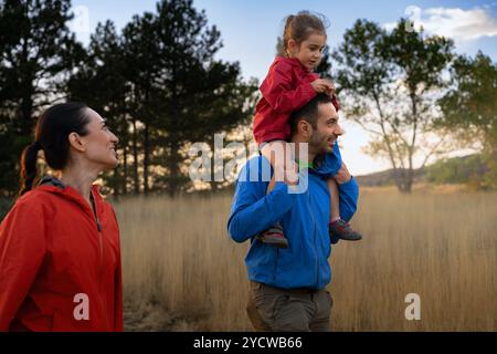Aktive Familie, die bei Sonnenuntergang Abenteuer in der Natur genießt. Vater trägt Tochter auf Schultern, während Mutter zusieht. Glückliche Momente beim Wandern Stockfoto