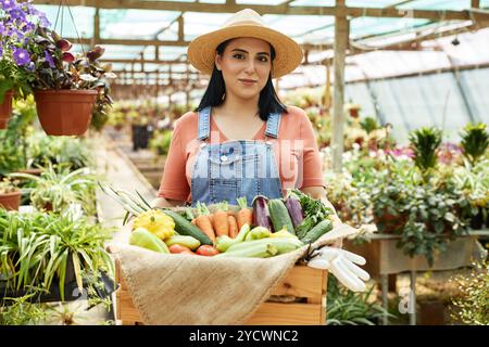 Porträt einer hispanischen Frau in Overall und Strohhut, die stolz frisch gepflücktes Gemüse in Gewächshäusern zeigt und lebendige Pflanzen und Blumen zeigt Stockfoto