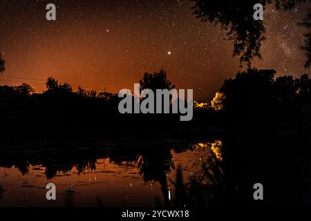 Sternenhimmel über dem Fluss und seine Reflexion im stillen Wasser. Der orangefarbene Himmel der Lichter und die Silhouette der Bäume spiegelten mich wider Stockfoto