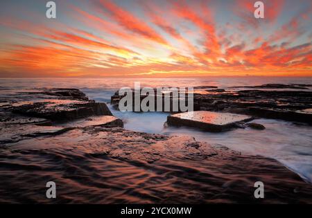 Roter Sonnenaufgangshimmel über dem Meer und erodierte Felskanäle und Einsiedlungen auf den dunklen, nassen Felsen. Lage North Narrabeen, nördliche Strände Sydney aus Stockfoto