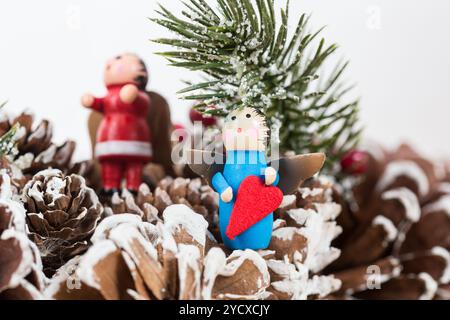 Weihnachtsschmuck mit einem kleinen Holzengel, der ein rotes Herz hält, vor einem festlichen Hintergrund aus Tannenzapfen und schneebedeckten Tannenzweigen Stockfoto