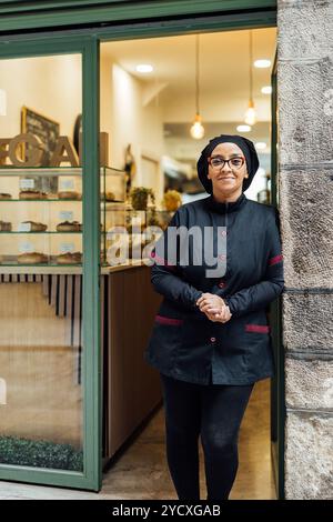 Eine selbstbewusste vegane Konditorin steht vor ihrer Bäckerei und lächelt in die Kamera, in einer professionellen Küchenuniform gekleidet, vor der einladenden Kulisse Stockfoto