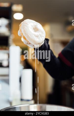 Eine Aufnahme eines frisch glasierten veganen Donuts, der über einer Schüssel gehalten wird, mit einer weißen Glasur, die auf dem Bild tropft, fängt die köstliche Textur und Süße ein Stockfoto