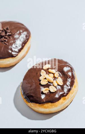 Zwei vegane Donuts mit glänzender Schokoladenglasur sitzen auf einer hellen Oberfläche, einer mit Erdnüssen, während der andere mit Schokoladenstreuseln belegt ist Stockfoto