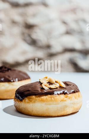 Zwei vegane Donuts mit glänzender Schokoladenglasur sitzen auf einer hellen Oberfläche, einer mit Erdnüssen, während der andere mit Schokoladenstreuseln belegt ist Stockfoto