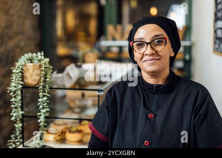 Ein lächelnder Bäcker steht selbstbewusst am Theken in einer gemütlichen veganen Bäckerei und präsentiert ein Angebot an frischem Gebäck Stockfoto