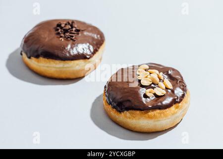 Zwei vegane Donuts mit glänzender Schokoladenglasur sitzen auf einer hellen Oberfläche, einer mit Erdnüssen, während der andere mit Schokoladenstreuseln belegt ist Stockfoto