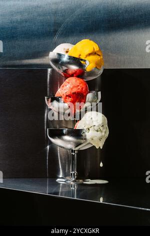 Verschiedene Eiskugeln, darunter Rot, Gelb und weiß, schmelzen verführerisch in einem schlanken Metallhalter vor einem sauberen, dunklen Hintergrund Stockfoto