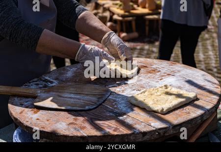 Aus nächster Nähe sehen Sie die Hände einer Person, die auf einem rustikalen Holzbrett auf einem Markt im Freien traditionelles georgisches Käsebrot, Khachapuri und Tonis Puri Formen Stockfoto