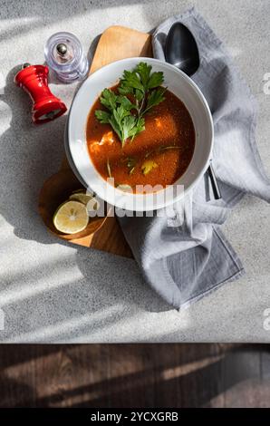 Blick von oben auf eine herzhafte Tomaten- und Kürbissuppe, garniert mit Käse, grünem Chili und frischer Petersilie, serviert in einer weißen Schüssel neben einem Glas Stockfoto