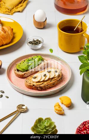 Ein lebhaftes Frühstück mit Avocado auf Getreidebrot, Erdnussbutter und Bananenscheiben auf Toast, Croissants, einer gelben Tasse Tee und frischem gar Stockfoto