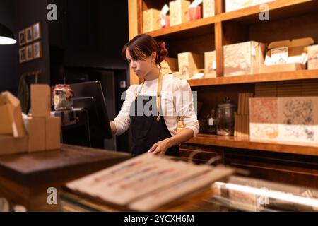 Eine junge Frau arbeitet fleißig an der Kasse in einem hübschen, gut sortierten Schokoladenladen, umgeben von verschiedenen wunderschön verpackten Pralinen und Betrügern Stockfoto