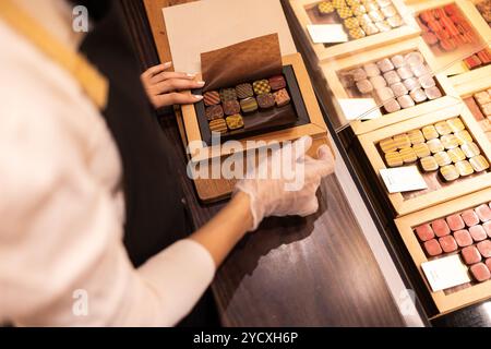 Arbeiter in weißem Hemd, schwarzer Schürze und Handschuhen, wählt feine Schokolade zum Verkauf in einem High-End-Schokoladengeschäft aus und präsentiert eine Vielzahl von bunten Gourmets Stockfoto