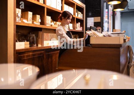 Eine Frau in einer eleganten Schürze arbeitet an der Kasse eines Gourmet-Schokoladengeschäfts, umgeben von Holzregalen mit verschiedenen handwerklichen Schokoladenprodukten Stockfoto