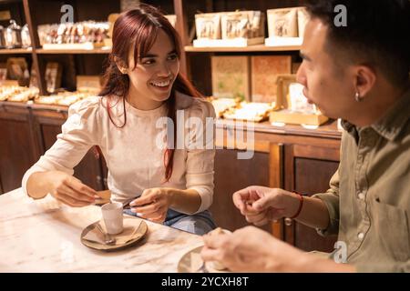 Eine junge Frau und ein Mann unterhalten sich bei einem Kaffee in einem gemütlichen Schokoladenladen, umgeben von Regalen voller Gourmet-Leckereien Stockfoto