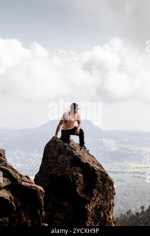 Ein junger Mann in einem Tank sitzt auf einem felsigen Gipfel am Cumbres del Ajusco und überblickt die weitläufige Wolkenlandschaft von Mexiko-Stadt Stockfoto