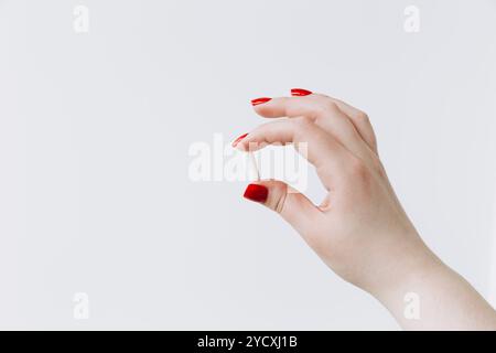 Weibliche Hand mit rot bemalten Nägeln hält eine weiße Heilpille sanft vor einem starken, isolierten weißen Hintergrund, der klare Fokus und minimalistische s Stockfoto