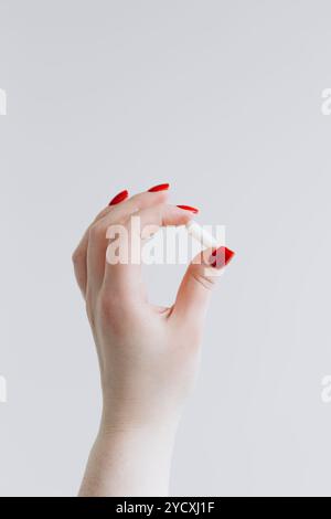 Eine Nahaufnahme einer weiblichen Hand, die einen feinen Griff auf einer kleinen weißen Pille zeigt. Die Hand ist mit elegantem rotem nagellack und einem glatten weißen bac versehen Stockfoto