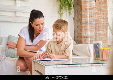 Eine fürsorgliche Mutter unterstützt ihren kleinen Sohn bei seiner Schularbeit in einer warmen, heimeligen Umgebung, umgeben von sanftem Tageslicht und Pflanzen im Innenbereich Stockfoto