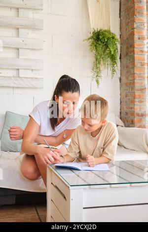 Eine fürsorgliche Mutter hilft ihrem kleinen Sohn bei der Schularbeit in einem gemütlichen, gut beleuchteten Zuhause, das sie bequem an einem modernen Glastisch umgeben von sitzen Stockfoto