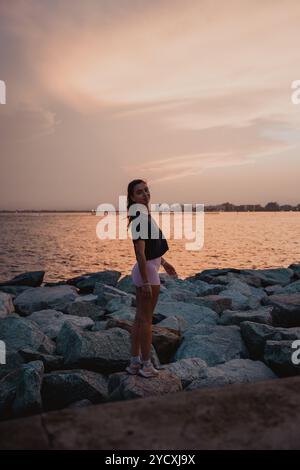 Eine junge Frau steht auf felsigem Gelände am Meer, in Laufkleidung vor einem dramatischen Abendhimmel und verkörpert das Wesen des Social Trail Running Stockfoto