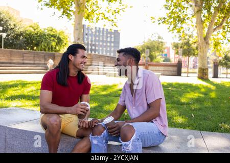 Multiethnisches schwules Paar, das im Freien in einem Stadtpark sitzt und verschiedene amerikanische lateinamerikanische und afroamerikanische Hintergründe repräsentiert Stockfoto