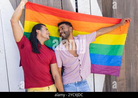 Fröhliches schwules Paar, bestehend aus einem amerikanischen Lateinamerikaner und einem Afroamerikaner, die die LGBTQIA+-Stolz-Flagge in einer Stadt halten und eine Vielfalt verkörpern Stockfoto