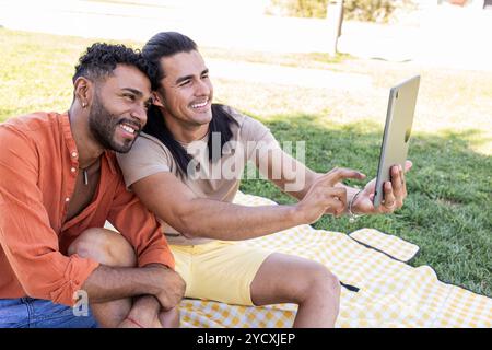 Ein fröhliches multiethnisches schwules Paar, ein Afroamerikaner und ein anderer Hispanic, sitzt auf einer Picknickdecke im Park, an dem sie einen Moment teilhaben Stockfoto