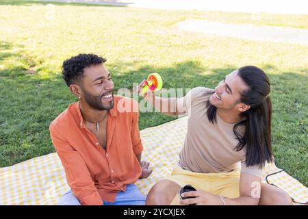 Ein fröhliches schwules Paar teilt einen fröhlichen Moment, indem es mit einem Handfan in einem Stadtpark spielt, was die Vielfalt der LGBTQIA+ Beziehungen widerspiegelt Stockfoto