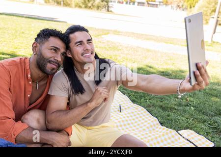 Ein fröhliches amerikanisches lateinamerikanisches und afroamerikanisches Schwulenpaar fängt an einem sonnigen Tag im Park ein Selfie ein und symbolisiert Liebe und LGBTQIA+ Stolz Stockfoto