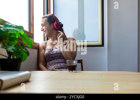 Eine fröhliche Transsexuelle hört Musik über rote Kopfhörer und spürt den Rhythmus, während sie zu Hause am Fenster sitzt. Genießen Sie einen sonnigen Morgen und einen Kaffee Stockfoto