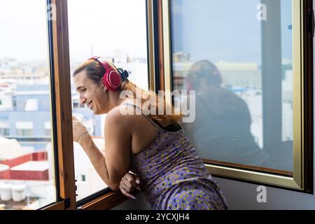 Eine fröhliche Transfrau in einem Blumenkleid trinkt Kaffee und hört Musik über rote Kopfhörer, blickt aus einem Fenster auf eine Stadtlandschaft und spiegelt po wider Stockfoto