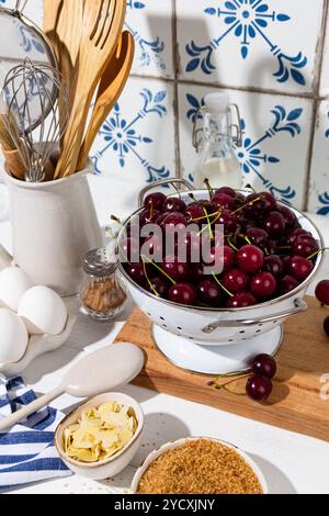Zutaten für Kirschkuchen, einschließlich Kirschen, Eier, Zimt und braunem Zucker, auf einem rustikalen Küchentisch mit Utensilien im Hintergrund. Stockfoto