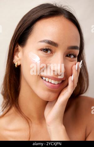 Porträt einer fröhlichen jungen Frau mit einem hauch Feuchtigkeitspflege auf der Wange, Hautpflegeprodukten und einem sanften Lächeln Stockfoto
