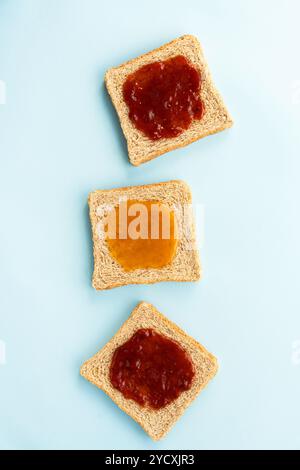 Drei Scheiben Weizenbrot mit verschiedenen Marmeladen, Erdbeeren und Pfirsichsorten, serviert auf hellblauem Hintergrund, perfekt für ein einfaches und doch delikates Erlebnis Stockfoto