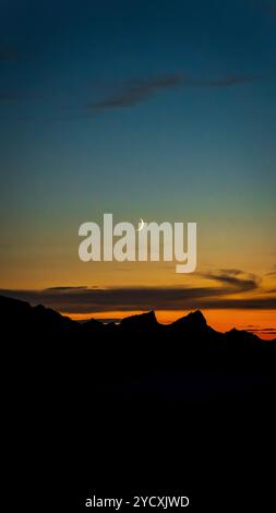 Ein atemberaubender Halbmond hängt am Abendhimmel über der Silhouette der Schweizer Alpen, mit einem farbenfrohen Sonnenuntergang, der den Horizont nahe dem GE-See erleuchtet Stockfoto