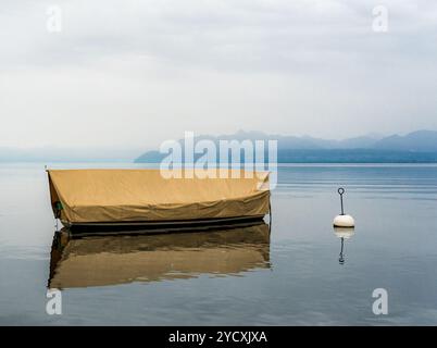 Eine ruhige Szene eines überdachten Bootes, das auf dem ruhigen Wasser des Genfer Sees in der Nähe der Ile de la Harpe in Rolle, Schweiz, schwimmt, mit Bergen im Hintergrund Stockfoto