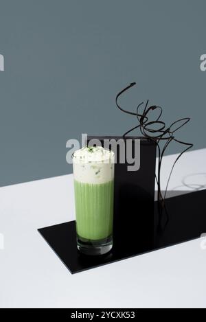 Ein erfrischendes Glas Eismatcha-Tee mit Schlagsahne, präsentiert auf einer eleganten schwarzen Plattform vor einem minimalistischen grauen Hintergrund. Stockfoto