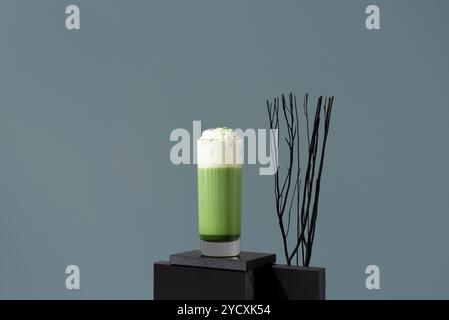 Ein gekühlter Matcha-Tee-Latte mit Schlagsahne, kunstvoll präsentiert auf einem minimalistischen Sockel mit stilistischen Zweigen im Hintergrund. Stockfoto