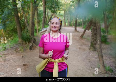 Reife Frau in rosa Hemd und Haarreif läuft mit einem Lächeln auf einem Waldweg, verkörpert Vitalität und aktives Altern Stockfoto