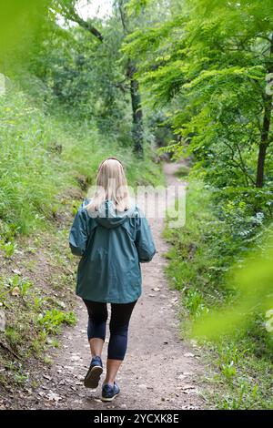 Eine reife Frau genießt einen friedlichen Spaziergang auf einem Waldweg, umgeben von üppigem Grün und natürlicher Schönheit Sie ist in bequeme Outdoor-Tuch gekleidet Stockfoto