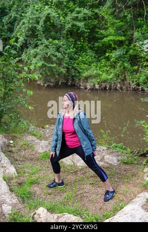 Eine aktive Seniorin macht während ihres Laufs auf einem bewaldeten Flussweg eine Pause, gekleidet in einem pinkfarbenen Oberteil, einer blauen Jacke und einer Leggings Stockfoto