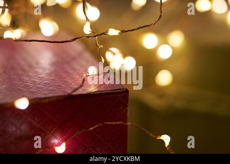 Ein Nahaufnahme-Bild mit einer roten Geschenkbox, die mit funkelnden Weihnachtslichtern geschmückt ist, vor einem Bokeh-Hintergrund aus goldenem Licht, das ein warmes Stockfoto