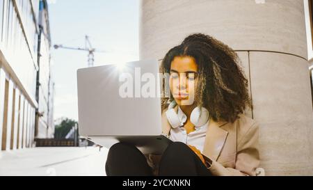 Eine junge Geschäftsfrau, die im Freien einen Laptop benutzt, sitzt auf dem Boden und hat Kopfhörer um den Hals. Sie arbeitet ferngesteuert in einer städtischen Umgebung Stockfoto