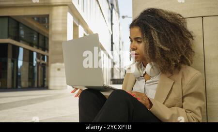 Die junge Geschäftsfrau sitzt draußen auf dem Boden und arbeitet mit ernstem Ausdruck an ihrem Laptop. Sie trägt einen Blazer und Kopfhörer Stockfoto