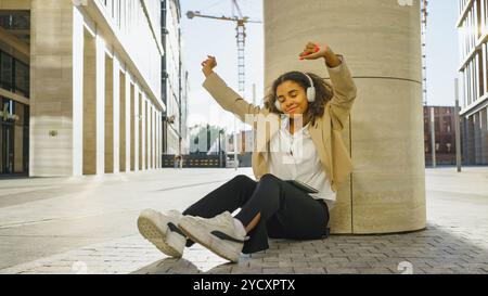 Die junge Geschäftsfrau sitzt draußen mit erhobenen Armen, hört Musik auf ihren Kopfhörern und genießt einen Moment der Entspannung Stockfoto