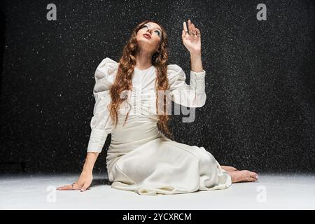 Eine anmutige junge Frau sitzt elegant in einem weißen Kleid, während der Schnee sanft um sie fällt. Stockfoto