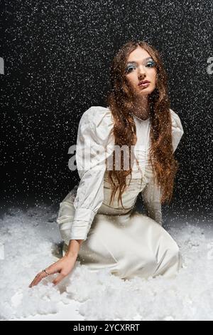 Eine junge Frau in eleganter weißer Kleidung kniet in einer verschneiten Umgebung, strahlt Pose und Schönheit aus. Stockfoto